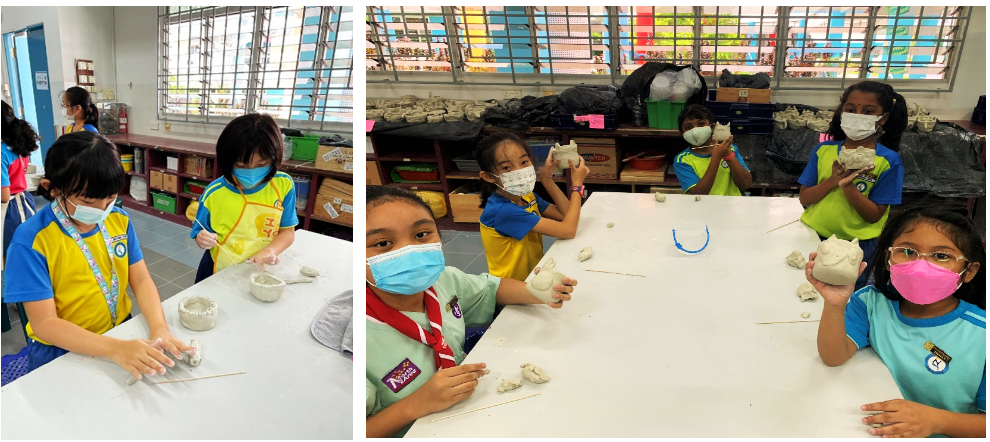 Children wearing face masks sit at tables in a classroom, shaping and holding white clay sculptures.